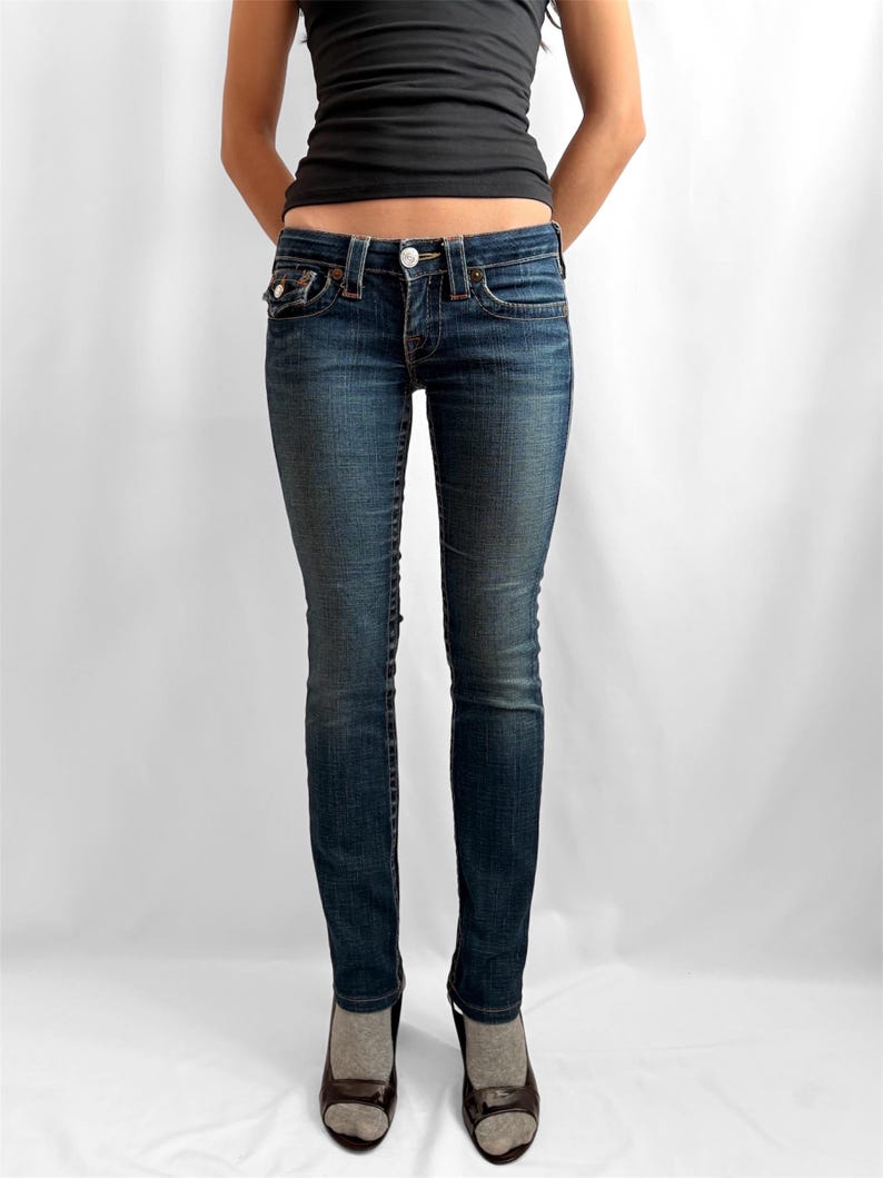 May include: Dark wash denim jeans with a classic five-pocket design. The jeans have a button and zipper closure, and are worn with a black tank top and dark shoes. The jeans are a slim fit.