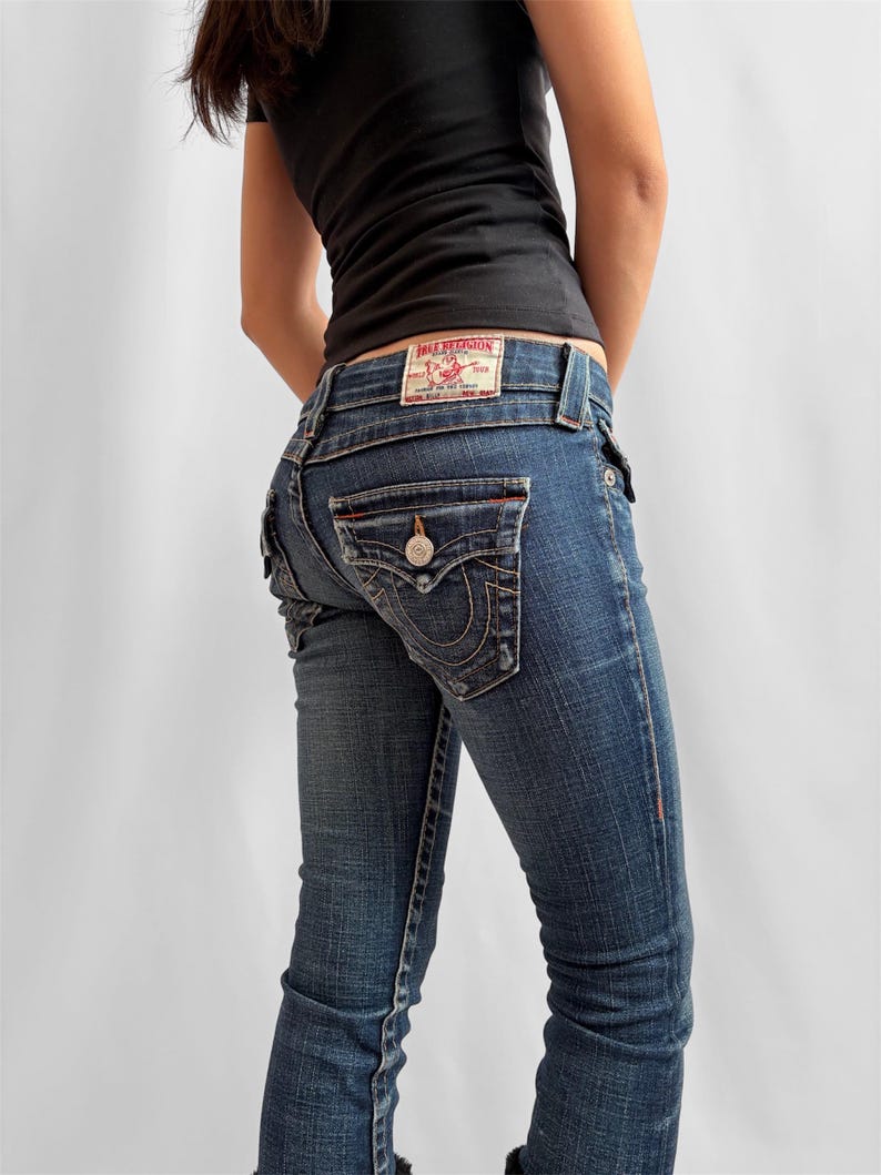 May include: Dark wash denim jeans with a button closure and back pocket stitching. The jeans have a red and white True Religion label on the back waistband. The jeans are a classic, everyday style.
