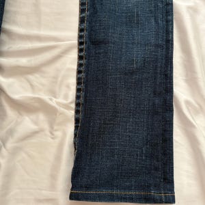 May include: Close-up of a section of dark blue denim jeans. The jeans have a straight leg cut with visible stitching in a contrasting yellow-orange thread along the seams and hem. The fabric has a textured appearance.