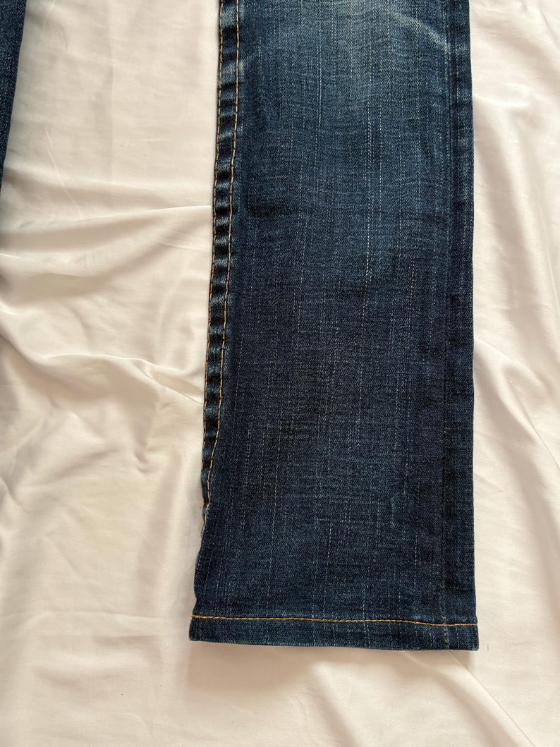 May include: Close-up of a section of dark blue denim jeans. The jeans have a straight leg cut with visible stitching in a contrasting yellow-orange thread along the seams and hem. The fabric has a textured appearance.