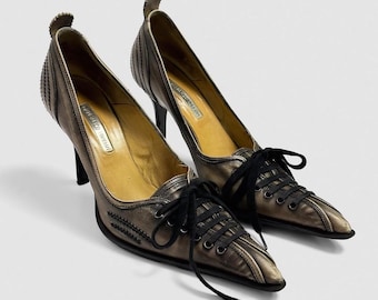 May include: A pair of pointed-toe court shoes in a metallic bronze finish with a distressed appearance. These shoes feature black lace-up closures on the vamp, black eyelets, and a high stiletto heel. The interior is lined in tan leather with a visible brand label.