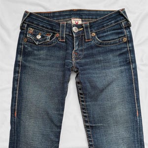 May include: Dark wash denim jeans with a classic five-pocket design. Features include a button and zipper closure, decorative stitching, and a branded metal button. The jeans have a faded appearance and are laid flat.