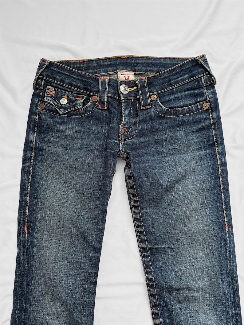 May include: Dark wash denim jeans with a classic five-pocket design. Features include a button and zipper closure, decorative stitching, and a branded metal button. The jeans have a faded appearance and are laid flat.