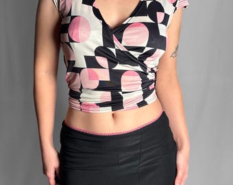May include: A short-sleeved wrap top featuring a geometric pattern of black, white, and pink circles and squares, paired with a black midi skirt that has a pink lace trim at the waist and a small pink bow detail at the front slit.