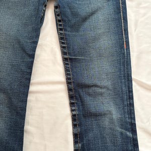 May include: Close-up of blue denim jeans. The jeans have a classic five-pocket design, with a button and zipper closure. The stitching is a contrasting color, and there are subtle fading details.