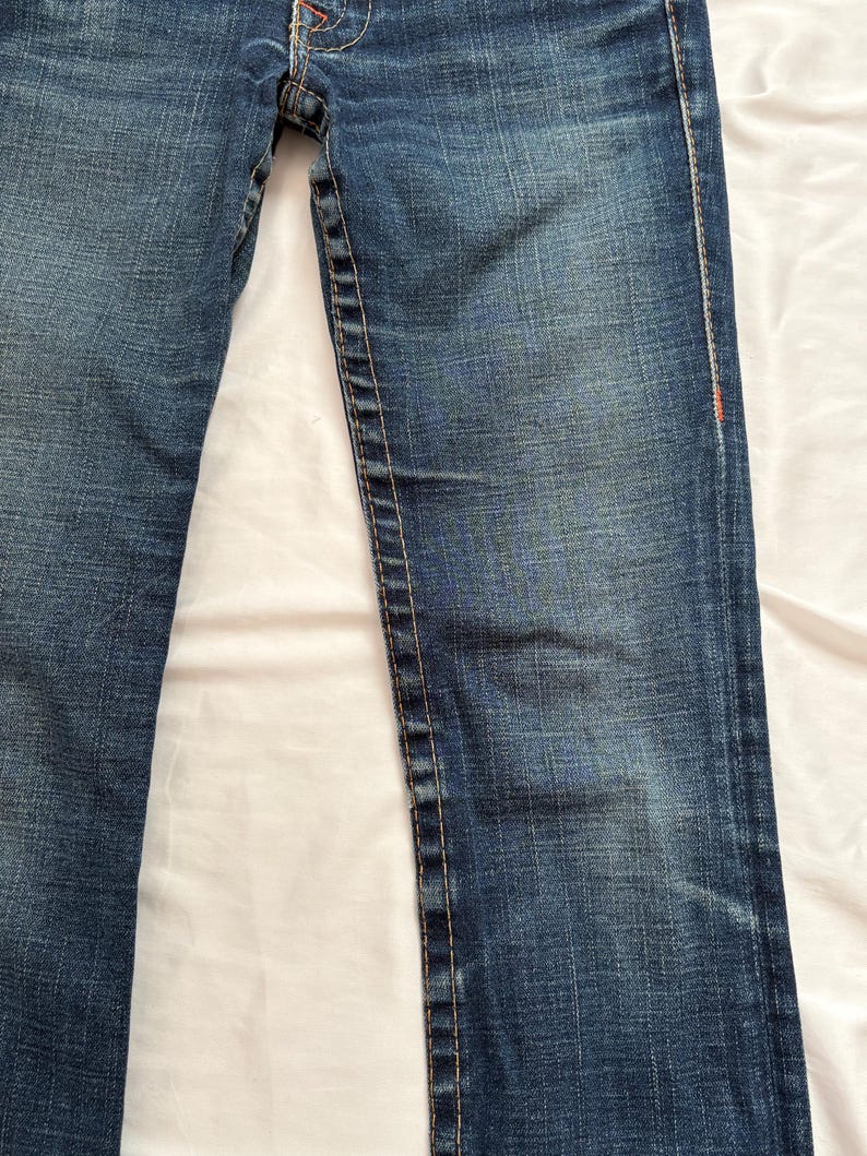 May include: Close-up of blue denim jeans. The jeans have a classic five-pocket design, with a button and zipper closure. The stitching is a contrasting color, and there are subtle fading details.