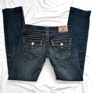May include: Dark wash denim jeans with a classic five-pocket design. Features decorative stitching on the back pockets and a branded leather patch. The jeans have a button and zipper closure.