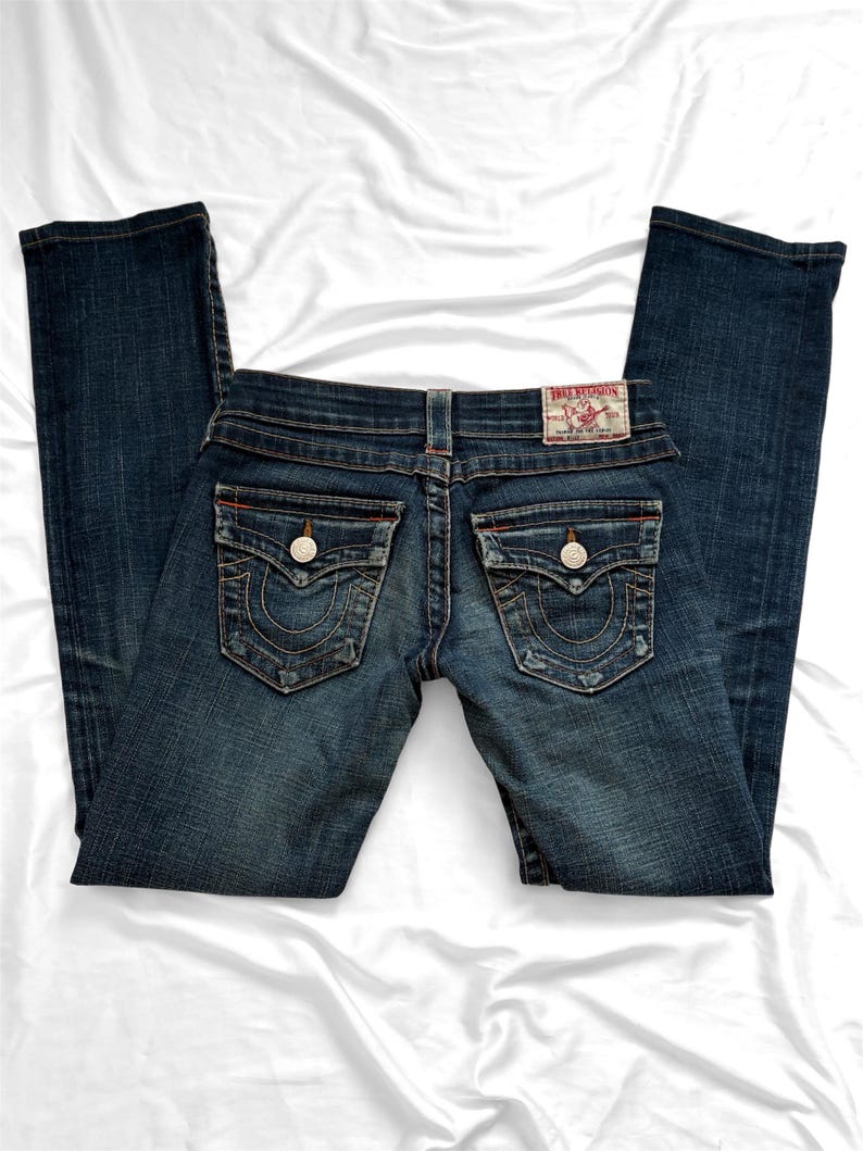 May include: Dark wash denim jeans with a classic five-pocket design. Features decorative stitching on the back pockets and a branded leather patch. The jeans have a button and zipper closure.