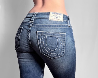 May include: Medium-wash blue denim jeans featuring white contrast stitching, back pockets with a signature horseshoe embroidery, and a white leather-look brand patch on the waistband that reads True Religion Brand Jeans World Tour.