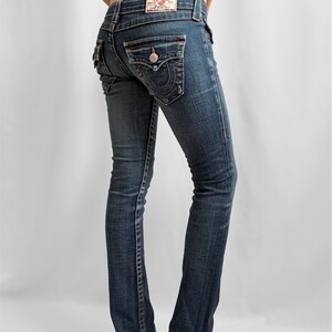 May include: Dark wash denim jeans with a bootcut leg. The jeans feature decorative stitching, a button-flap back pocket, and a red and white label. The jeans are worn with high-heeled shoes.