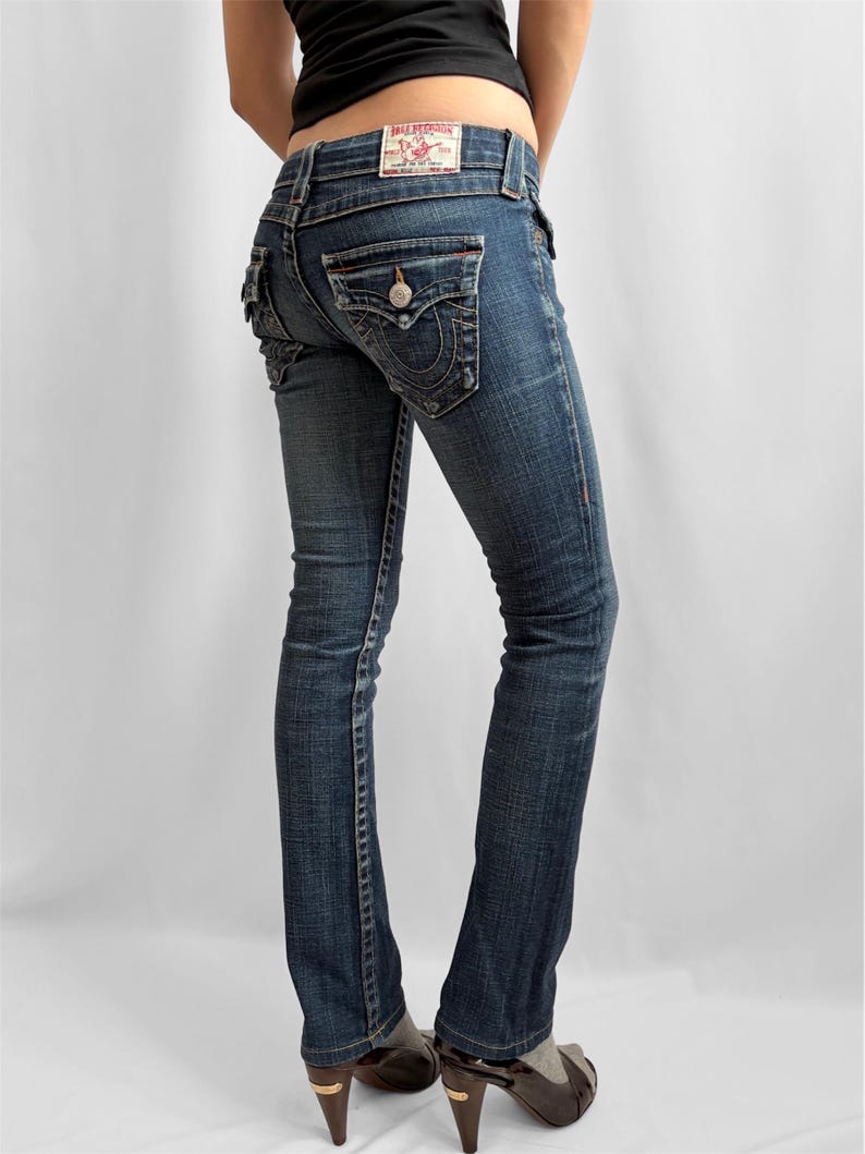 May include: Dark wash denim jeans with a bootcut leg. The jeans feature decorative stitching, a button-flap back pocket, and a red and white label. The jeans are worn with high-heeled shoes.