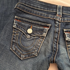 May include: Close-up of dark wash denim jeans. The jeans feature a classic five-pocket design with a distinctive horseshoe-shaped pocket stitch. The back pocket has a silver button. The stitching is in a contrasting gold color, with red accents.