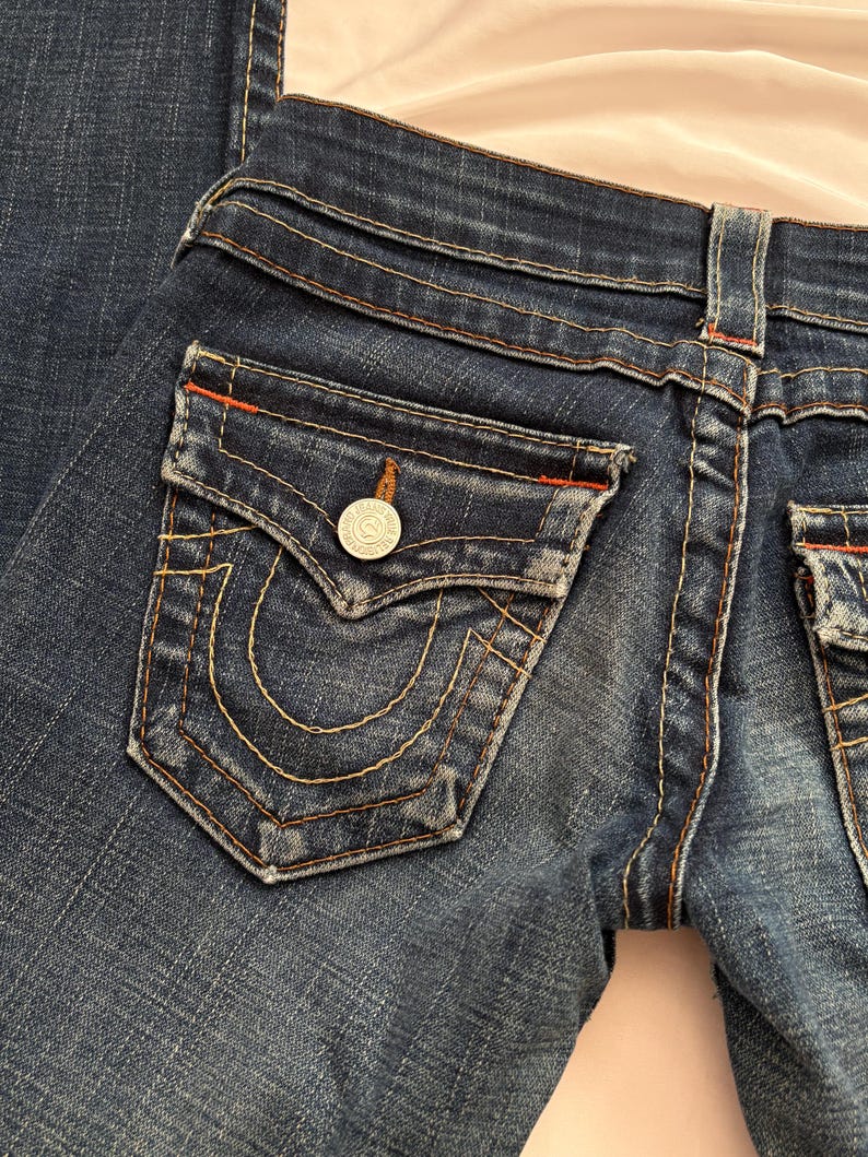 May include: Close-up of dark wash denim jeans. The jeans feature a classic five-pocket design with a distinctive horseshoe-shaped pocket stitch. The back pocket has a silver button. The stitching is in a contrasting gold color, with red accents.