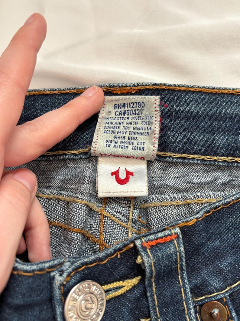 May include: Close-up of dark blue denim jeans with visible stitching and a red and white tag. The tag reads "RN#112790 CA#3042" and washing instructions. A small white tag with a red horseshoe logo is also visible.