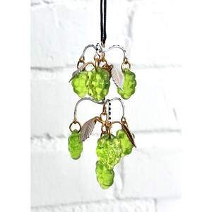 May include: A green glass berry cluster pendant with gold and white accents hangs from a black cord. The berries are clustered in groups of three and five, and the pendant is suspended from a delicate wire frame.
