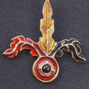 May include: A gold, red, and black metal pin with a stylized leaf design and a round center with a black stone.