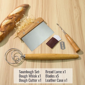 May include: A sourdough baking set with a wooden dough whisk, a dough cutter, a bread lame with a leather case and five blades. The set is on a wooden surface with a loaf of bread and wood shavings.