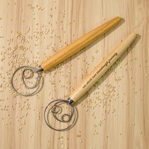 May include: Two wooden dough whisks with metal loops. The whisk on the right has the text "we start our sourdough journey" engraved on the handle.