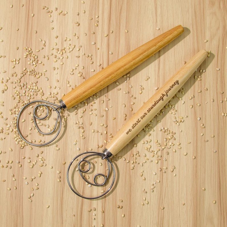 May include: Two wooden dough whisks with metal loops. The whisk on the right has the text "we start our sourdough journey" engraved on the handle.