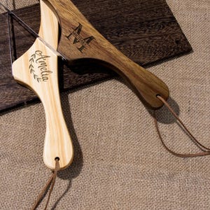 Puede incluir: Dos tablas de cortar de madera con correas de cuero. Una tabla es de color marr&oacute;n claro y est&aacute; grabada con el nombre "Amelia" y una corona de laurel. La otra tabla es de color marr&oacute;n oscuro y est&aacute; grabada con el nombre "Michael" y un monograma.