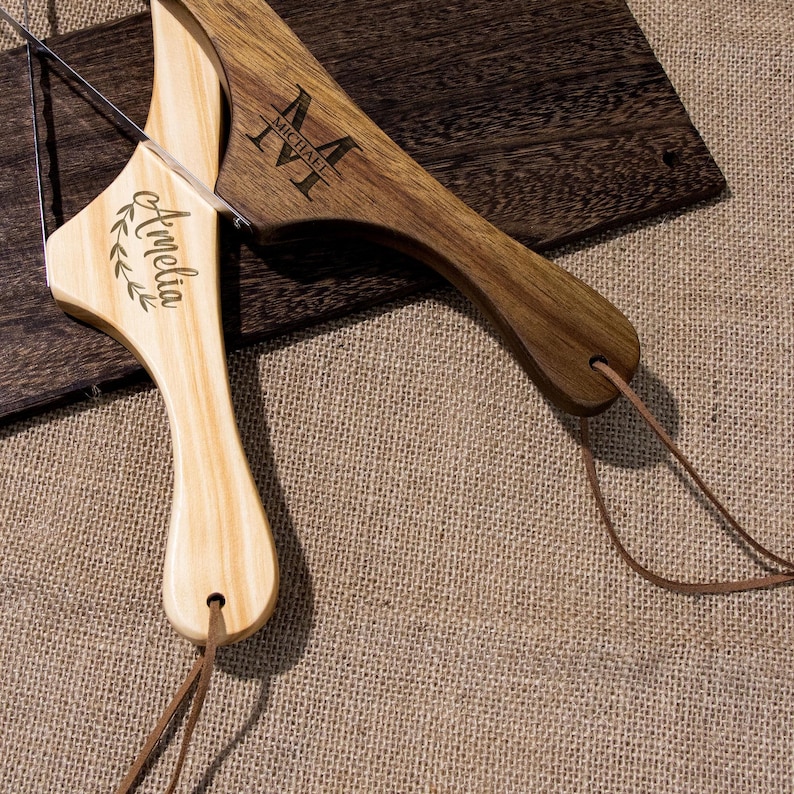 Puede incluir: Dos tablas de cortar de madera con correas de cuero. Una tabla es de color marr&oacute;n claro y est&aacute; grabada con el nombre "Amelia" y una corona de laurel. La otra tabla es de color marr&oacute;n oscuro y est&aacute; grabada con el nombre "Michael" y un monograma.