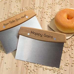 Pode incluir: Duas espátulas de bancada de aço inoxidável com cabos de madeira, uma com a inscrição "Happy Bake Love Again" e a outra com "Happy Gains". Um bagel castanho-dourado repousa sobre uma pequena tábua de madeira, rodeado por grãos, numa superfície de madeira.