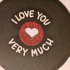 Puede incluir: Una tarjeta de felicitaci&oacute;n con un dise&ntilde;o de disco de vinilo negro. El centro del disco es un c&iacute;rculo de purpurina roja con un recorte en forma de coraz&oacute;n. El texto blanco dice "I LOVE YOU VERY MUCH" alrededor del disco.