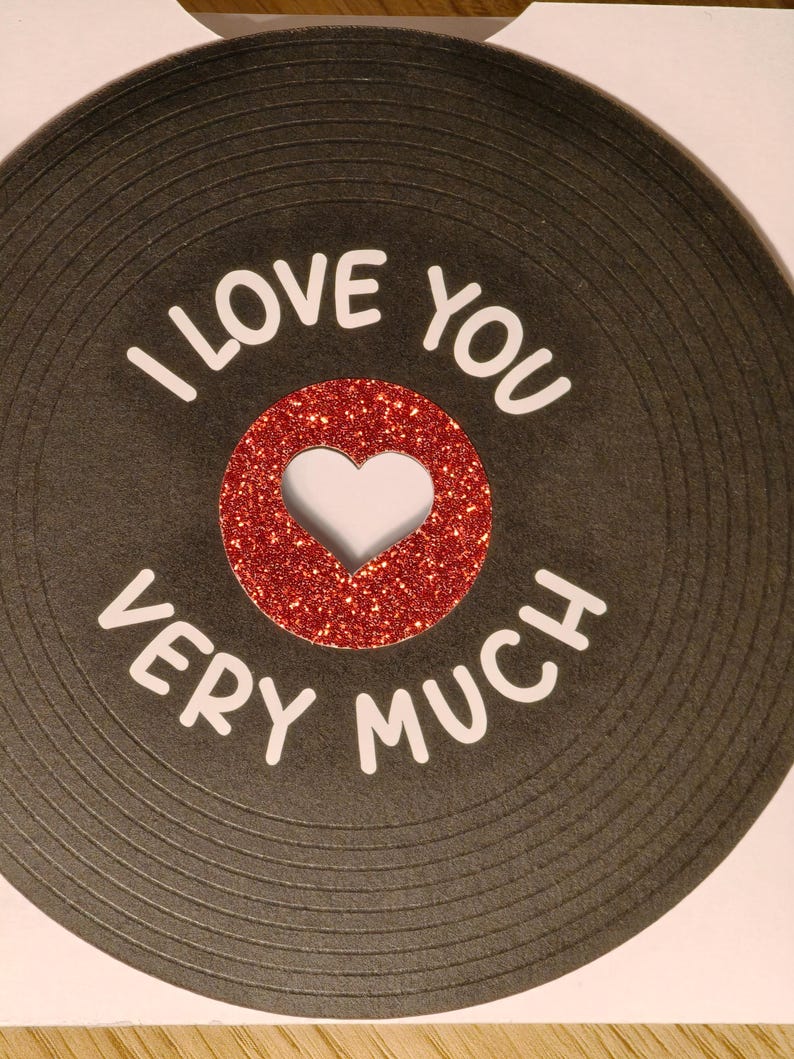 Puede incluir: Una tarjeta de felicitaci&oacute;n con un dise&ntilde;o de disco de vinilo negro. El centro del disco es un c&iacute;rculo de purpurina roja con un recorte en forma de coraz&oacute;n. El texto blanco dice "I LOVE YOU VERY MUCH" alrededor del disco.