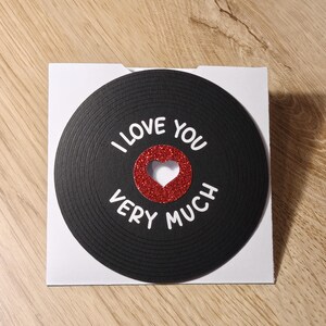 Puede incluir: Tarjeta de felicitaci&oacute;n con un dise&ntilde;o de disco de vinilo negro. Las palabras "I LOVE YOU VERY MUCH" est&aacute;n escritas en blanco. Un coraz&oacute;n rojo brillante est&aacute; en el centro. La tarjeta est&aacute; sobre un fondo blanco.