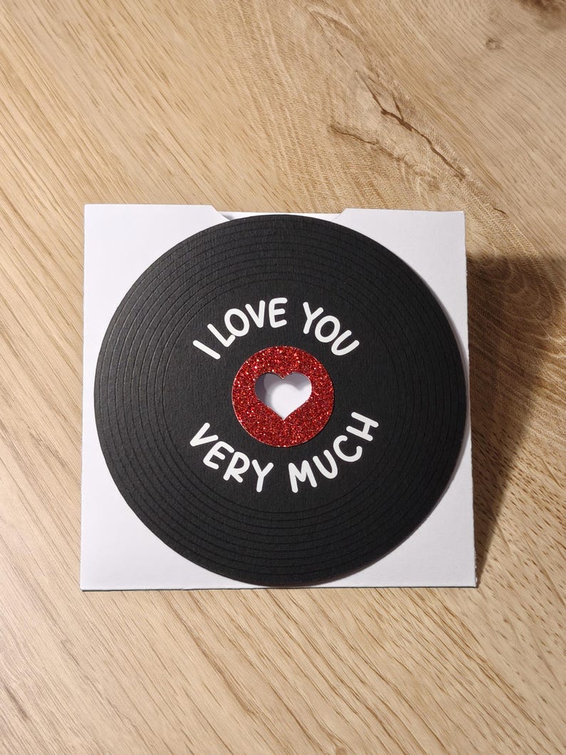 Puede incluir: Tarjeta de felicitaci&oacute;n con un dise&ntilde;o de disco de vinilo negro. Las palabras "I LOVE YOU VERY MUCH" est&aacute;n escritas en blanco. Un coraz&oacute;n rojo brillante est&aacute; en el centro. La tarjeta est&aacute; sobre un fondo blanco.