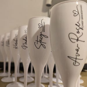 May include: A row of white champagne flutes arranged in a line, each featuring black cursive text and a small heart icon. The visible names include Ava Rose, Stacy, Nadia, Karah, and others, with titles like Chief Bridesmaid and Bridesmaid.