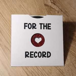 Puede incluir: Una tarjeta blanca con el texto "FOR THE RECORD" en negro. Una forma de coraz&oacute;n con purpurina roja est&aacute; recortada de la tarjeta. La tarjeta est&aacute; sobre una superficie de madera.
