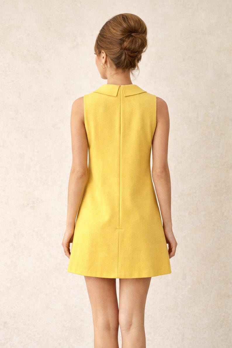 May include: A sleeveless, yellow dress with a collared neckline. The dress has a simple, A-line silhouette and falls above the knees. The model's hair is styled in a bouffant updo.