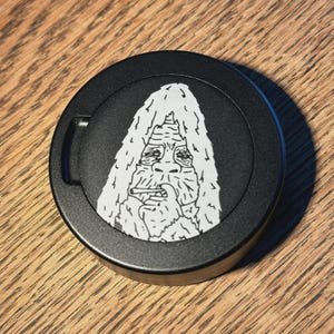 Puede incluir: Un recipiente redondo negro con un gráfico blanco de un Bigfoot fumando un cigarrillo. La ilustración de Bigfoot está detallada con pelo blanco y una expresión seria. El recipiente tiene un pequeño asa curvada.