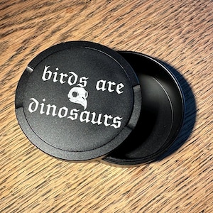 Può includere: Contenitore nero e rotondo con la scritta "birds are dinosaurs" in bianco, in carattere gotico. Un teschio di uccello bianco è tra le parole. Il contenitore è aperto, rivelando l'interno.