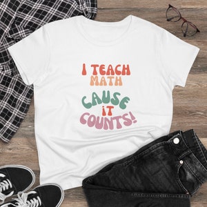 May include: White t-shirt with the text "I TEACH MATH CAUSE IT COUNTS!" in retro-style lettering. The text is in shades of red, orange, green, and pink. The shirt is displayed on a wooden surface with black jeans, black and white sneakers, and eyeglasses.