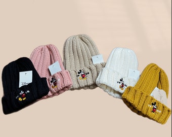 May include: Five knit beanies in different colors, black, pink, beige, white, and yellow, with a Mickey Mouse embroidered on the front. Each beanie has a small tag attached.