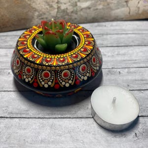May include: A black candle holder with a mandala design painted in red, yellow, and gold. The holder has a small green succulent plant in the center and a white tea light candle.