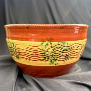 May include: A ceramic bowl featuring a glossy finish with horizontal bands of deep red and a central yellow section. The yellow band is decorated with embossed wavy lines and scattered green speckles, creating a textured appearance.
