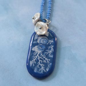 May include: A necklace featuring a blue beaded chain and a rectangular pendant with a glossy finish. The pendant displays a white pattern of seashells and coral on a dark blue background. Two small, white, flower-shaped beads are attached.