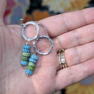 May include: A pair of dangle earrings featuring hammered silver-tone circular hoops. Each earring has a vertical stack of four small, ribbed glass beads in alternating light blue and lime green colors, suspended from the silver-tone metal hoops.