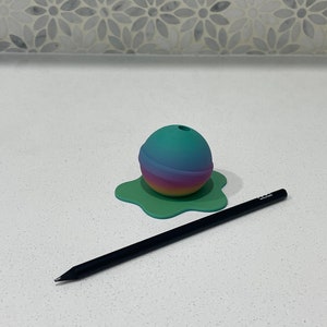 Op de afbeelding: Een turquoise, roze en gele ombre potloodhouder in de vorm van een bol met een gat aan de bovenkant. De houder staat op een groene, splatter-vormige basis.
