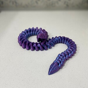 Op de afbeelding: Een 3D-geprint slangen speelgoed in paars en blauw met een gesegmenteerd lichaam. De slang is opgerold in een spiraalvorm.