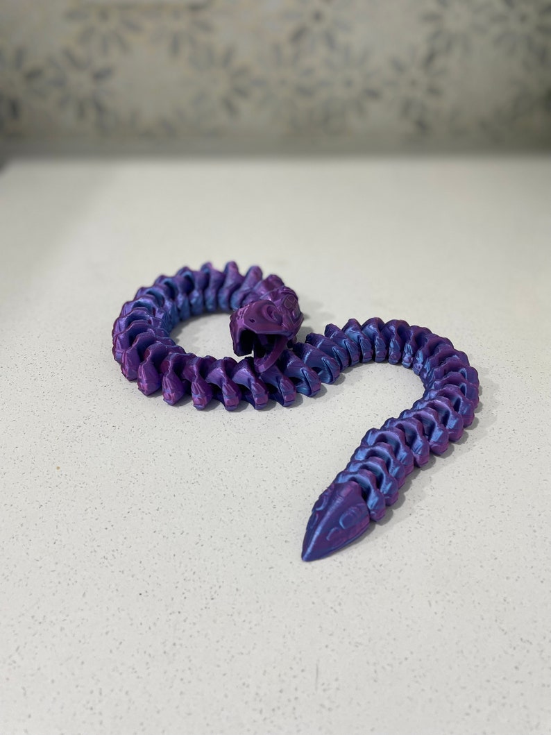 Op de afbeelding: Een 3D-geprint slangen speelgoed in paars en blauw met een gesegmenteerd lichaam. De slang is opgerold in een spiraalvorm.