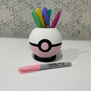 Op de afbeelding: Een witte en roze Pokeball-vormige bureau-organizer met een zwarte knop aan de voorkant. De organizer is gevuld met kleurrijke pennen en stiften. Een roze en grijze Sharpie permanente marker ligt op de tafel.