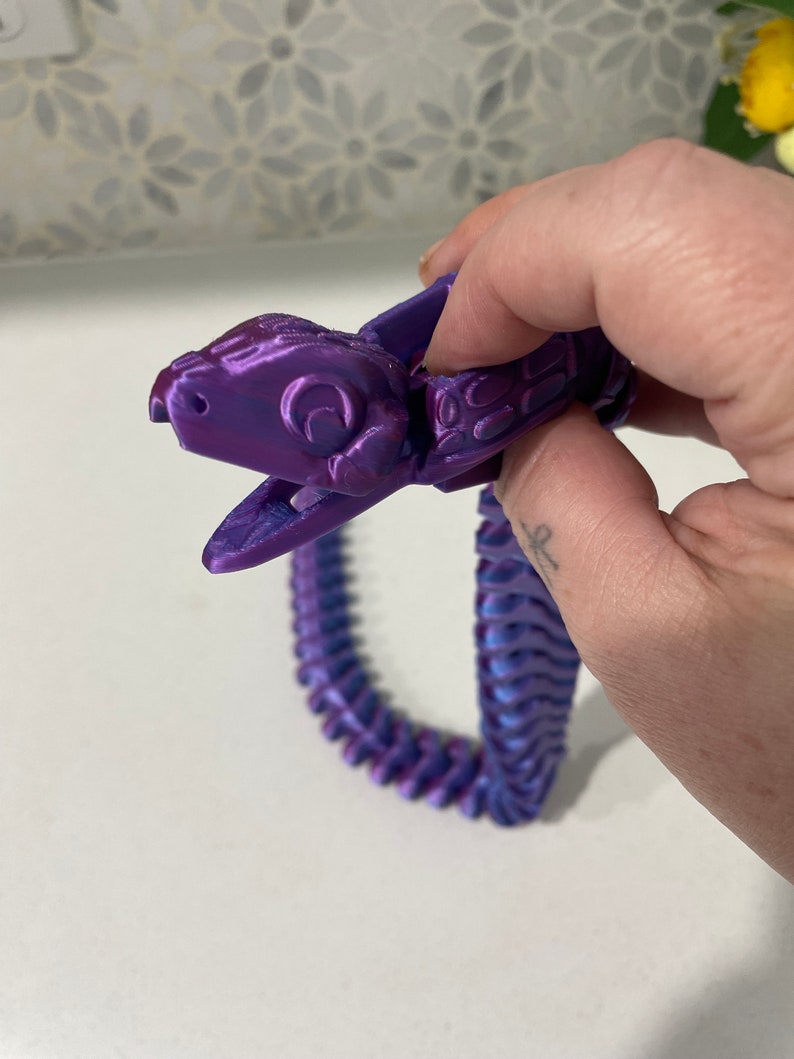 Op de afbeelding: Een 3D-geprint slangen speelgoed in paars en blauw. De slang is gesegmenteerd en heeft een gedetailleerd hoofd met een textuur.