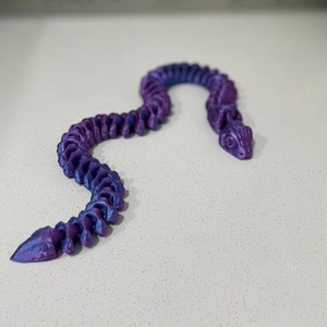 Op de afbeelding: Een paarse en blauwe iriserende plastic slangachtige armband met een gesegmenteerd ontwerp.