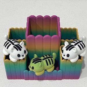 Op de afbeelding: Een regenboogkleurige bureau-organizer met vier vakken. De organizer heeft een groen gestreept kattenbeeld in het middelste vak en twee witte gestreepte kattenbeeldjes in de zijvakken.