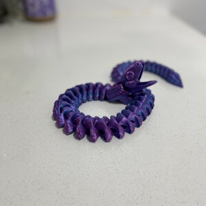 Op de afbeelding: Een slangen-vormig speelgoed van plastic in een iriserende paarse en blauwe kleur. Het speelgoed is opgerold in een cirkel met de kop omhoog.