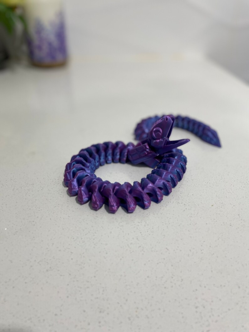 Op de afbeelding: Een slangen-vormig speelgoed van plastic in een iriserende paarse en blauwe kleur. Het speelgoed is opgerold in een cirkel met de kop omhoog.